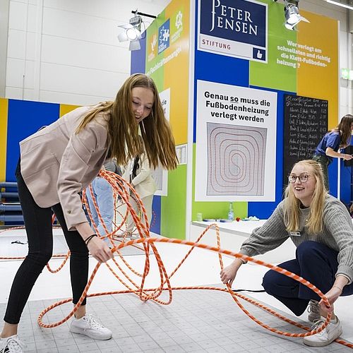 Zwei junge Frauen simulieren eine Verlegung einer Fußbodenheizung. 