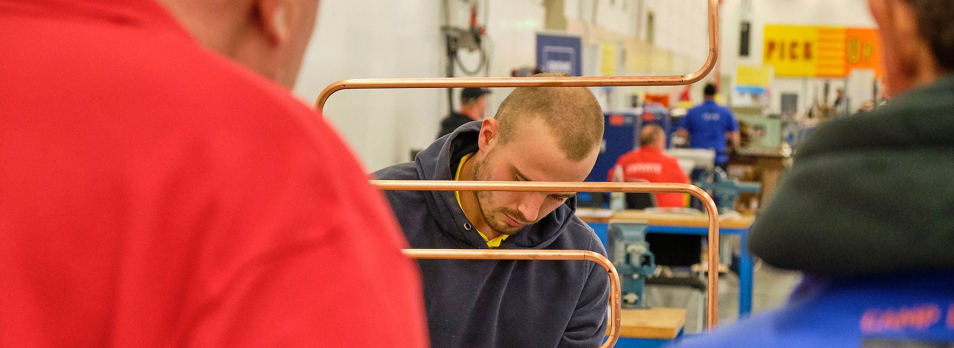 In der Mitte des Bildes arbeitet ein Teilnehmer an seinem Projekt für die Meisterschaft der Klempnerei. Man sieht ein gebogenes Kupferrohr. Vor ihm stehen mit dem Rücken zum Betrachtenden zwei Jury-Mitglieder.