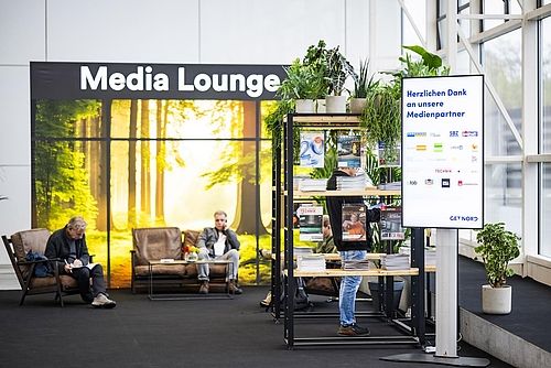 Blick von Eingang Mitte auf die Media Lounge. Mehrere gemütliche Sessel stehen vor einer Wand mit Naturdruck und dem Schriftzug Media Lounge. Daneben stehen einige Regale mit Zeitschriften der Medienpartner.