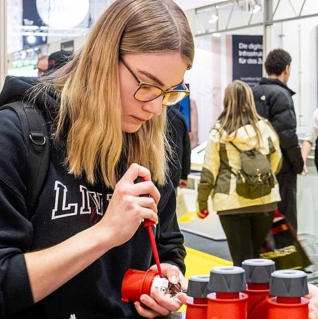 Eine junge Frau auf der linken Bildseite öffnet konzentriert mit einem Schraubenschlüssel ein kleines Gerät. Ein Mann auf der rechten Seite unterstützt sie dabei.