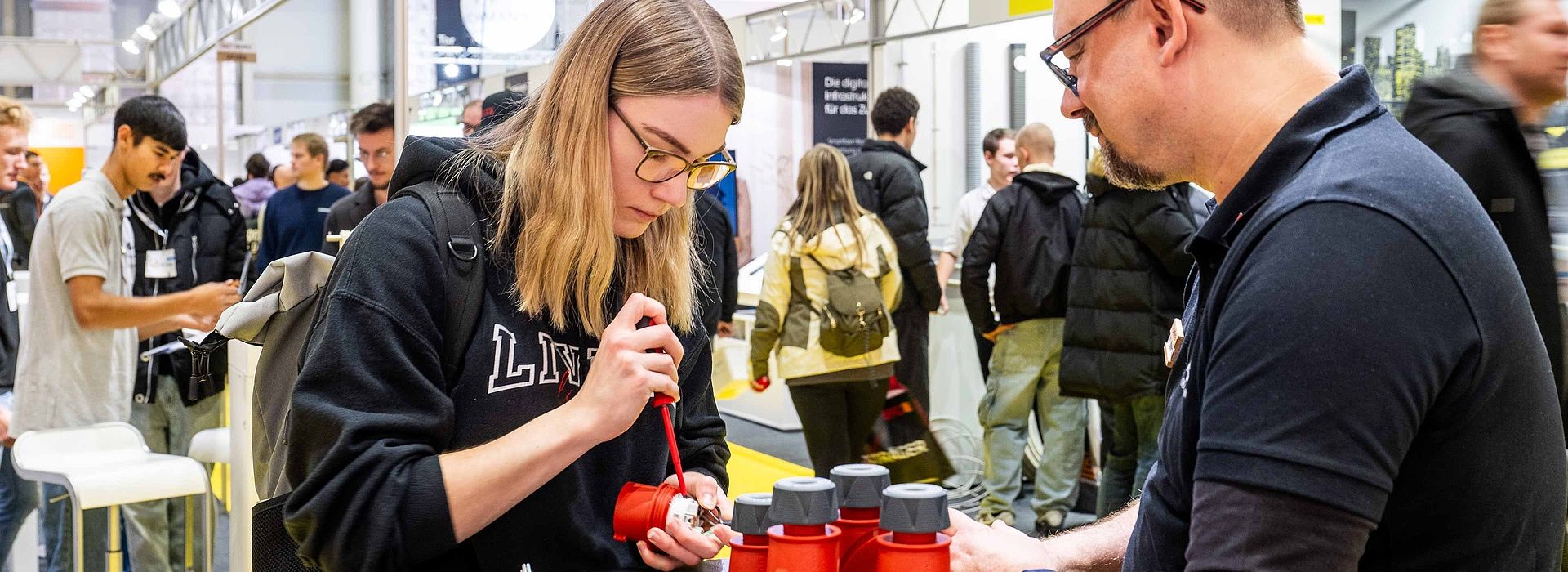 Eine junge Frau auf der linken Bildseite öffnet konzentriert mit einem Schraubenschlüssel ein kleines Gerät. Ein Mann auf der rechten Seite unterstützt sie dabei.