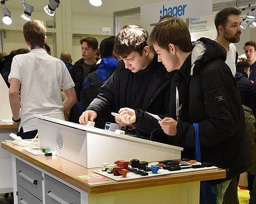 Auf dem Bild sind zwei junge Männer zu sehen, die an einem Messestand technische Bauteile untersuchen. Im Hintergrund sind weitere Personen in einer belebten Ausstellungshalle zu erkennen.