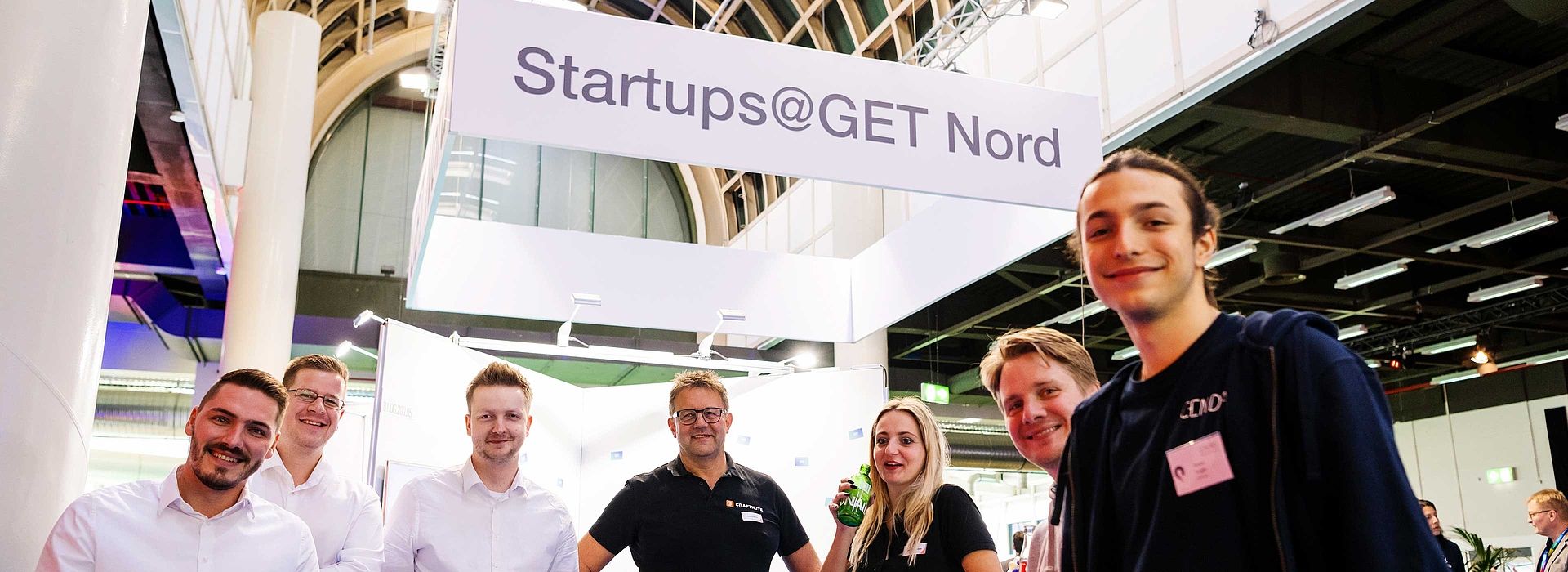 Sieben Personen schauen in die Kamera. Im Hintergrund hängt ein Schild mit Startups@GET NORD.