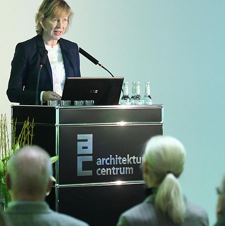 Ein Redner steht am Podest eines Architekturforums und hält eine Präsentation vor dem Publikum